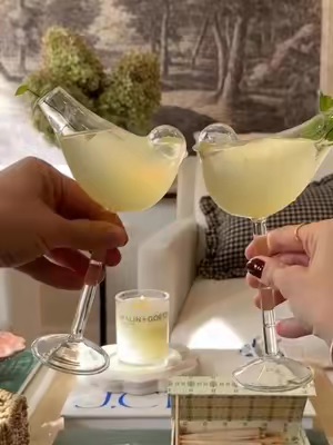 Two hands hold up bird-shaped glasses filled with a pale yellow beverage, clinking them in a toast. A small candle sits between them, suggesting a cozy and celebratory atmosphere.