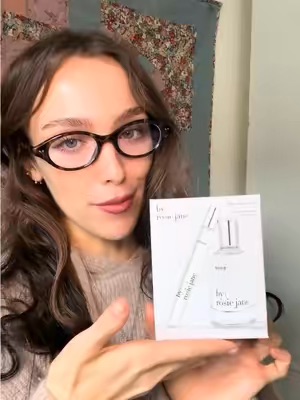 A woman with glasses is holding up a small white perfume sample card towards the camera in a casual indoor setting with a floral-patterned quilt in the background. This appears to be a UGC handheld demo showcasing beauty products.