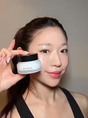 A young woman with smooth, glowing skin is smiling while holding up a jar of skincare moisturizer directly to the camera in a close-up view. This is a UGC handheld demo showcasing a Korean skincare product.