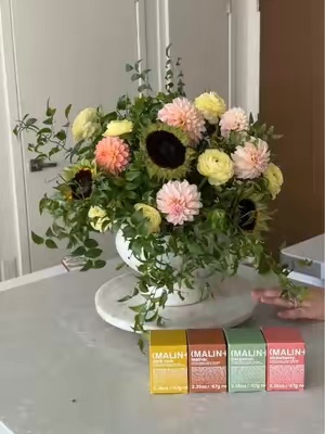 A beautiful floral arrangement sits on a marble cake stand, with four small boxes of MALIN+GOETZ votives arranged in front of it. The scene evokes a sense of sophisticated home decor and sensory experience.
