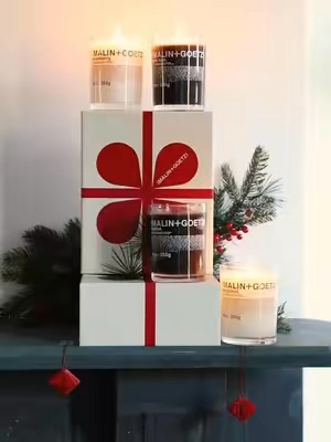 A festive display of MALIN+GOETZ candles is presented as a gift set, adorned with red ribbon and surrounded by evergreen branches, evoking a sense of holiday warmth and luxury.