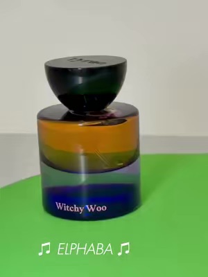 The image shows a colorful glass fragrance bottle labeled 'Witchy Woo' placed on a bright green surface with a text overlay, resembling a simple studio product shot promoting a themed perfume.