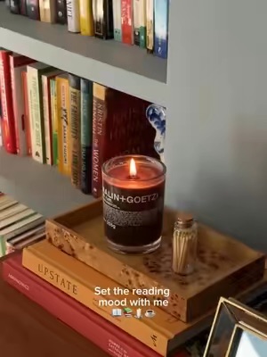 A MALIN+GOETZ Dark Rum candle is lit and placed on a tray with matches, surrounded by books on a shelf, creating a cozy reading atmosphere.