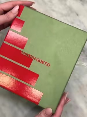 A close-up shot of a Malin+Goetz branded gift box, held by hands, highlighting its elegant and ready-to-gift presentation.