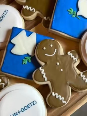 The image features a close-up of assorted decorated cookies, including gingerbread men and ones with blue icing and white dove designs, arranged on a wooden surface in a flat lay style.